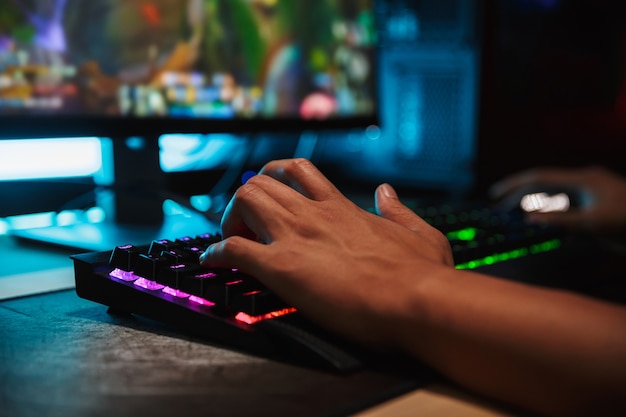 Mãos de um jovem jogador jogando videogame no computador em um quarto escuro, usando um teclado colorido com luz de fundo