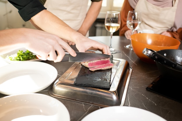 Mãos de um jovem treinador de cozinha cortando um pedaço de carne grelhada ou defumada em um quadro especial ao lado da mesa com dois estagiários de pé perto