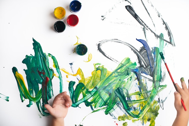 Mãos de um menino pintando com aquarelas na folha de papel branco. Garotinho com um pincel e tintas.