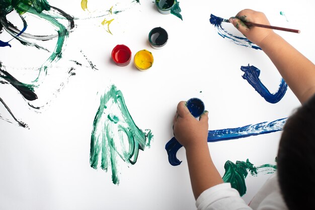 Mãos de um menino pintando com aquarelas na folha de papel branco.