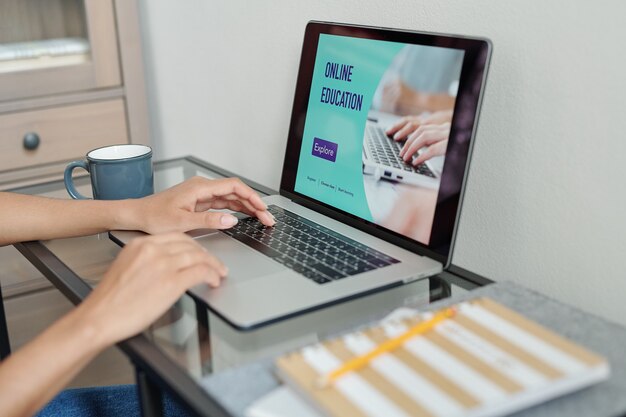 Mãos de uma aluna contemporânea apertando botões do teclado enquanto está sentada à pequena mesa em frente ao laptop durante a aula online em casa