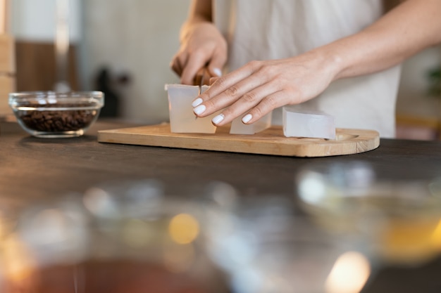 Mãos de uma jovem com uma faca cortando um pedaço de massa de sabão duro na placa de madeira enquanto fazem produtos cosméticos naturais em casa