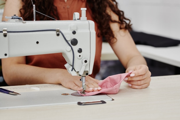 Mãos de uma jovem costureira contemporânea costurando ombreiras cor-de-rosa enquanto está sentada à máquina e terminando o trabalho em um novo item