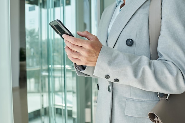 Mãos de uma jovem empresária elegante em terno cinza mandando mensagens no celular