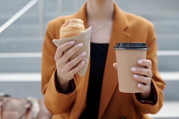 Mãos de uma jovem empresária moderna segurando croissant fresco e uma xícara de café