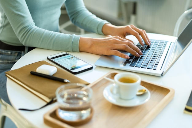 Mãos de uma jovem empresária ou freelancer em roupa casual tocando as teclas do teclado do laptop enquanto trabalha em rede à mesa no café