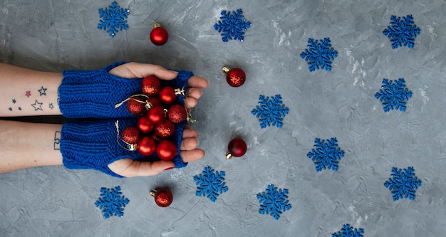 Mãos de uma jovem mulher bonita em luvas azuis. Ela segura bolas vermelhas de Natal. Perto estão flocos de neve azuis e decorações vermelhas na árvore de Natal.