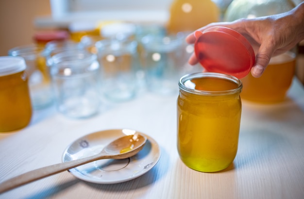 Mãos de uma mulher desconhecida fecham potes de vidro transparente com mel dourado puro e doce com tampa de borracha, em pé sobre uma grande mesa de madeira branca e ao lado de um pequeno pires com uma colher de pau