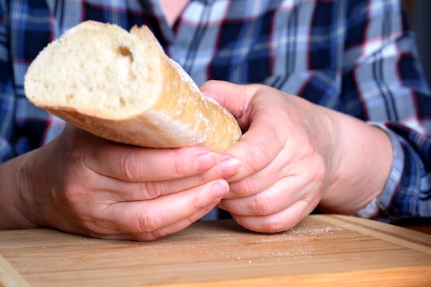 Mãos de uma mulher idosa segurando baguete de pão de trigo branco fatiado na cozinha em uma mesa marrom sem rosto fechado