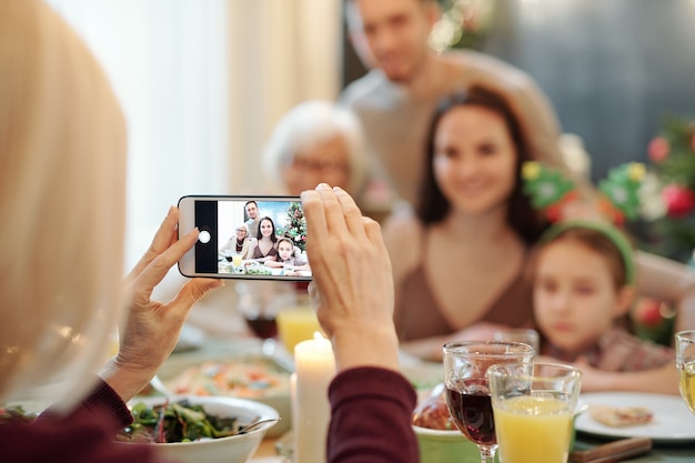 Mãos de uma mulher madura segurando um smartphone enquanto tira uma foto do jovem casal, sua filha e a avó no jantar