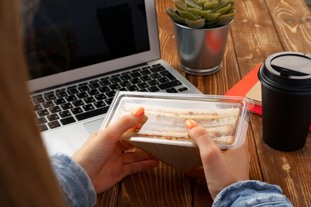 Mãos de uma mulher segurando um sanduíche acima da mesa de trabalho com um laptop