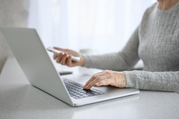 Mãos de uma mulher sênior trabalham em um laptop com telefone, usando tecnologia moderna na vida cotidiana. compras online