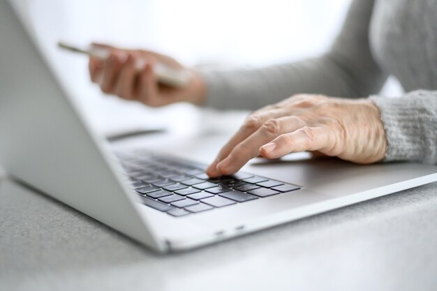 Mãos de uma mulher sênior trabalham em um laptop com um telefone, usando tecnologia moderna na vida cotidiana.