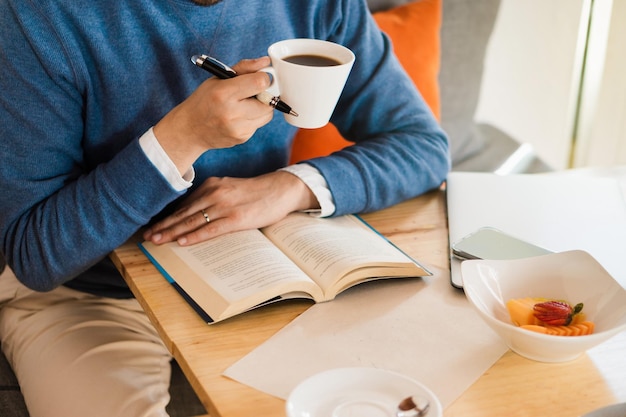 Mãos de uma pessoa irreconhecível estudando com um livro e segurando um café.