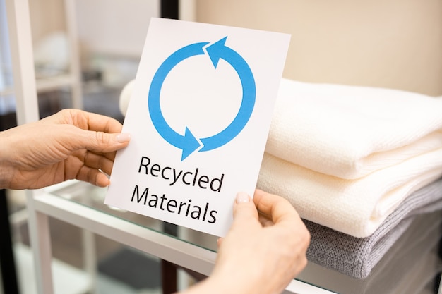 Mãos de uma vendedora segurando um cartão dizendo sobre materiais reciclados contra uma pilha de vestidos quentes de lã dobrados na prateleira