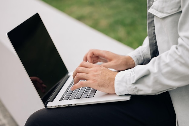 Mãos de usuário masculino digitando no teclado do laptop sentadas no banco, empresário étnico de raça mista, estudante hipster