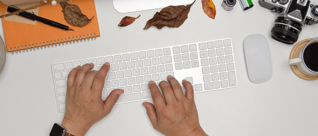 Mãos digitando no teclado do computador sem fio na mesa de escritório branca com câmera, artigos de papelaria e decorações