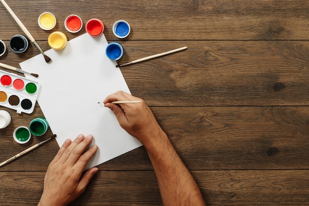 Mãos do artista closeup, segurando um pincel sobre uma folha de papel em branco perto de latas abertas de tintas coloridas