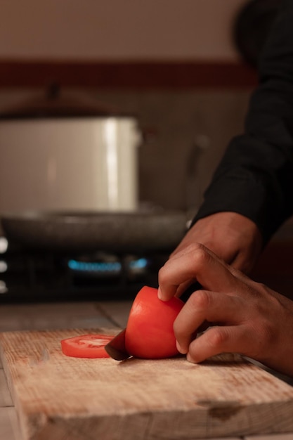 Mãos do chef masculino cortando um tomate vermelho fresco na placa de madeira rústica com uma faca na cozinha