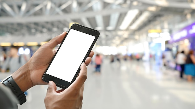 Mãos do homem inteligente, segurando um smartphone preto recortado com tela em branco branca sobre o aeroporto internacional lotado como turva