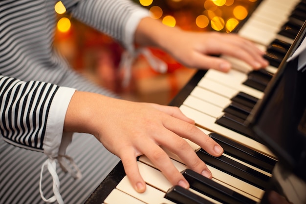 Mãos do jovem pianista no piano, close-up, luzes de Natal no fundo. Conceito de música e decoração de férias de inverno.