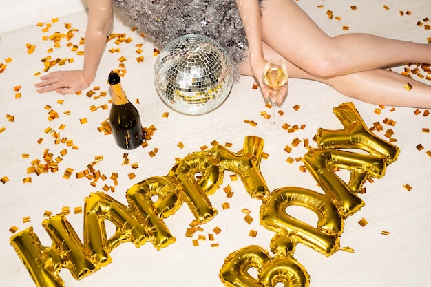 Mãos e pernas finas de uma jovem mulher glamourosa com taça de champanhe, comemorando o aniversário enquanto está sentada no chão com confete dourado