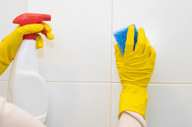 Mãos em luvas de borracha amarela lavando os azulejos do banheiro.