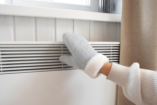 Mãos em luvas de inverno aquecendo sobre aquecedor elétrico mulher congelada vestindo luvas de malha congelando para o frio do inverno desconforto passando tempo em casa menina aquecendo as mãos no radiador moderno