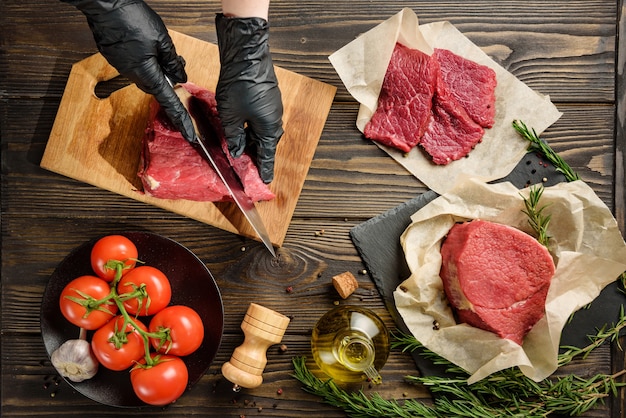 Mãos em luvas de proteção pretas cortam a carne contra a mesa de outros produtos alimentícios. Ingredientes para cozinhar bifes.