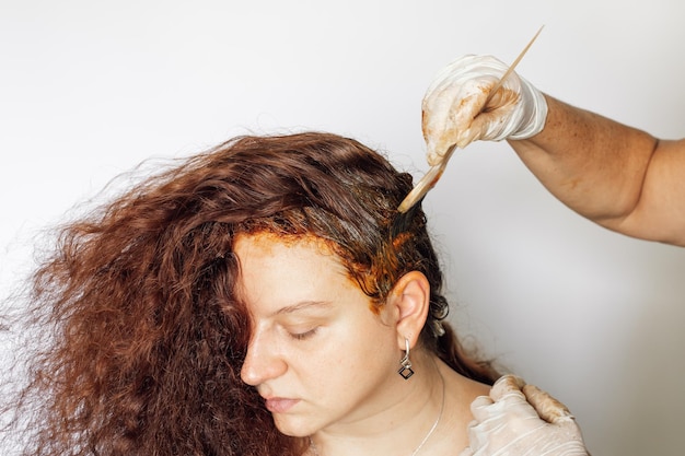 Mãos em luvas finas de látex aplicando alguma substância de tingimento no cabelo da mulher com escova plástica branca no fundo branco Coloração do cabelo em casa Livrando-se dos cabelos grisalhos Antiidade