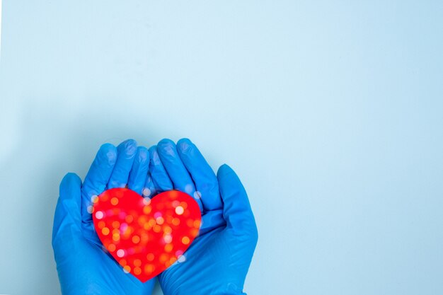 Mãos em luvas médicas azuis segurar um coração vermelho em uma mesa azul, cópia espaço, vista superior. O efeito bokeh é como um floco de neve.