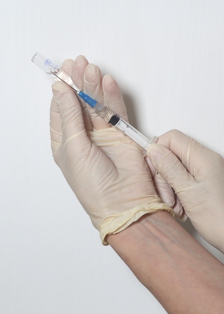 Mãos em luvas médicas enchem uma seringa com remédio de uma ampola fechada em um fundo branco