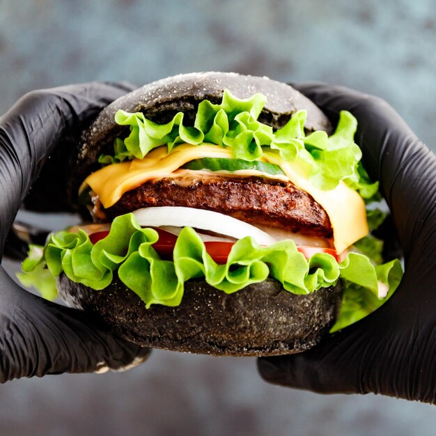 Mãos em luvas pretas seguram um grande hambúrguer preto com queijo de carne de mármore e legumes frescos vista aproximada