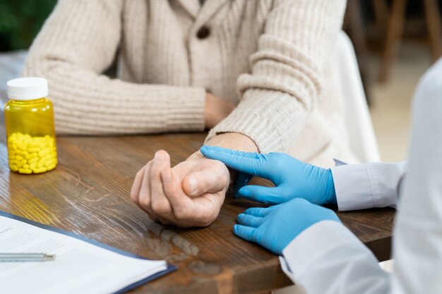 Mãos enluvadas de médico de clínica geral em jaleco branco tocando o pulso de paciente idoso do sexo masculino à mesa durante o check-up médico em casa