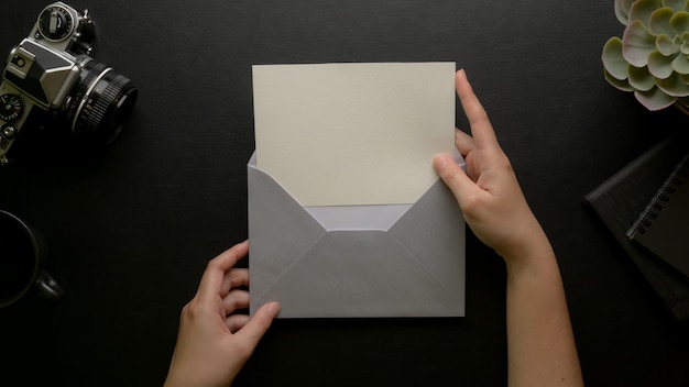 Mãos femininas abrir cartão com envelope cinza na mesa de escritório escuro com câmera e decorações