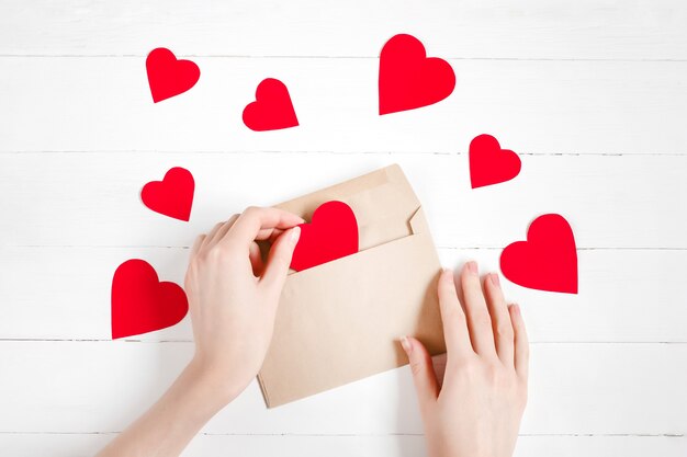 Mãos femininas colocar corações vermelhos em um envelope de felicitações em uma madeira branca