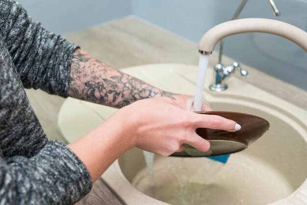 Mãos femininas com tatuagem lavam a placa de cerâmica sob água corrente da torneira