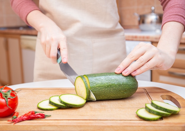 Mãos femininas cortam com uma faca uma jovem abobrinha sazonal em fatias sobre uma tábua de madeira. o método de preparação de vegetais. produtos sazonais, colheita. nutrição saudável, dieta alimentar.