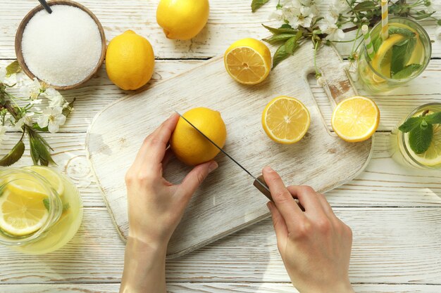 Mãos femininas cortam limão para fazer limonada em fundo branco de madeira