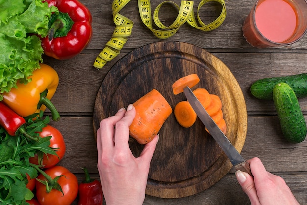 Mãos femininas cortando cenoura na mesa, vista superior. sobre a mesa folhas de alface, pimenta, um copo de suco de tomate, uma tábua de madeira e uma faca