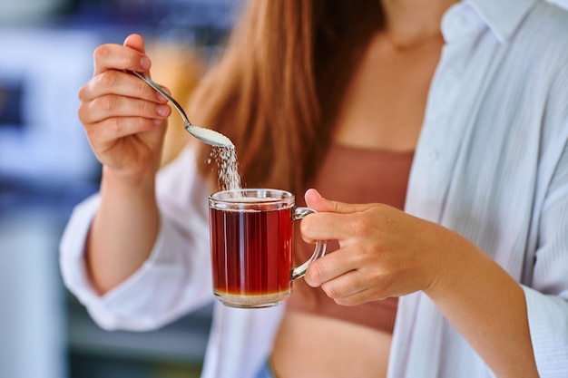 Mãos femininas derramando colher de açúcar em copo de vidro com chá preto quente