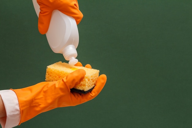 Mãos femininas em luva protetora laranja com frasco de detergente e esponja sobre o fundo verde. Conceito de lavagem e limpeza.