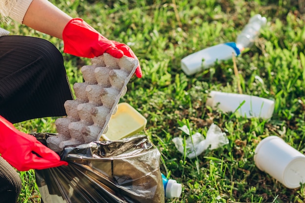 Mãos femininas em luvas de borracha vermelha. Mulher recolhe lixo no saco. Voluntários limpam lixo no parque de verão. Bela mulher progressiva fazendo um esforço para ajudar o meio ambiente