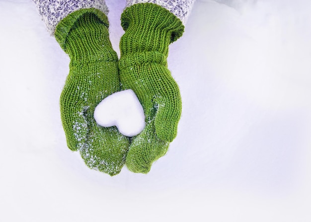 Mãos femininas em luvas de malha com coração de neve no dia de inverno, fundo do dia de São Valentim