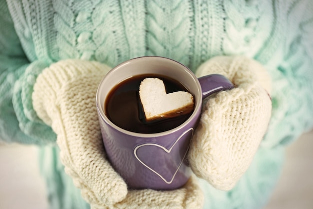 Mãos femininas em luvas quentes segurando uma xícara de café quente com marshmallow de coração close-up