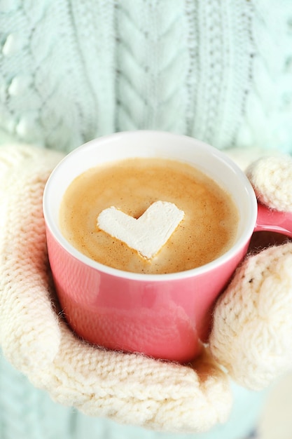 Mãos femininas em luvas quentes segurando uma xícara de cappuccino quente com marshmallow de coração close-up