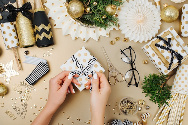 Mãos femininas embrulhando presentes na mesa com fundo de decoração de natal nas cores douradas e pretas. camada plana, vista superior