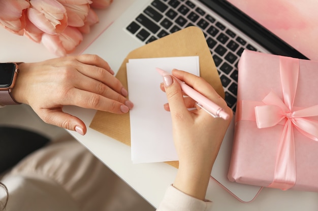 Mãos femininas escrevendo desejos no último papel para o dia dos namorados ou aniversário com caixa de presente e laptop na mesa, vista superior.
