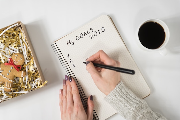 Mãos femininas escrevendo meus objetivos 2020 em um caderno. caneca de café e homem-biscoito, vista superior