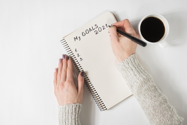Mãos femininas escrevendo My Goals 2021 em um caderno. Caneca de café na mesa, vista superior.